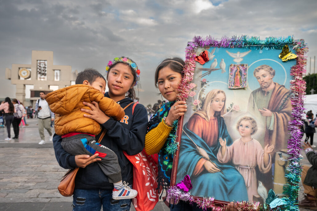 Dos jóvenes frente a la Basílica de la Virgen de Guadalupe en México. Una carga en brazos a un niño y la otra carga una imagen de la Sagrada Familia.